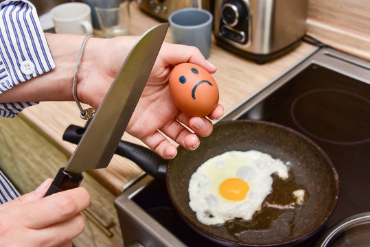 woman in the kitchen in front of the pan holds a knife and breaks a sad smiling egg