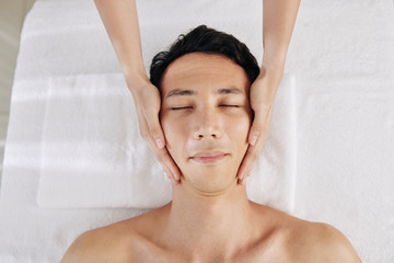 Mixed-race man enjoying rejuvenating face massage, masseuse stroking slowly up the sides of his...