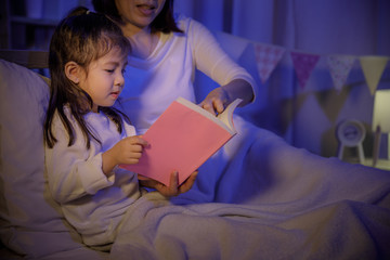 Mom reading a book with her daughter on bed in a dark bedroom at night before sleep, Parenthood and...