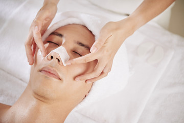 Fototapeta na wymiar Close-up image of beautician putting sticky nose strip on face of young man lying on bed in spa salon