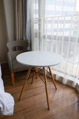 Modern White round table with wooden legs and gray armchair near window in hotel room. Concept for minimal style in living room or relaxing area.