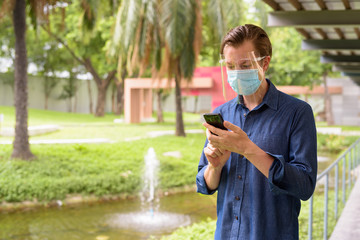 Young man with mask and face shield for protection from corona virus outbreak using phone at the park