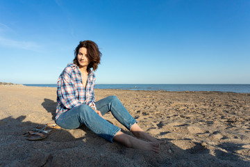 Fototapeta na wymiar A cheerful dark-haired woman smiles, sitting on the beach, enjoys the bright sun on a summer day. Concept of summer holidays at sea and live style