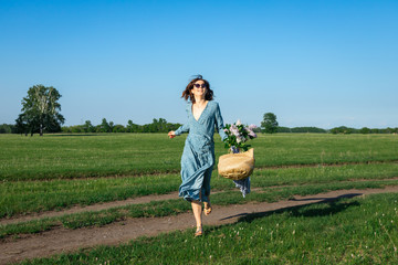 A cheerful dark-haired woman smiles, walks along the green field dressed in a romantic blue dress on the smell, wicker bag with a bouquet  on a summer day. 