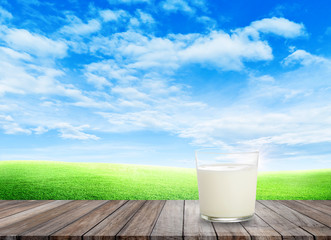 White milk in glass on wooden table with green grass field and little hill in background.