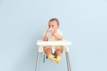 Cute little baby with bottle sitting on feeding chair against color background
