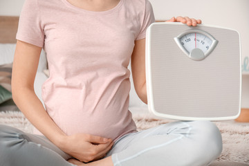 Young pregnant woman with measuring scales in bedroom