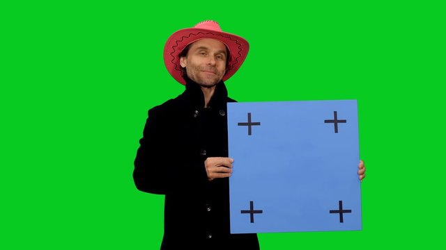 Stylish cheerful man in black coat and pink cawboy hat advertises product via blank blue board against green screen background, Chroma key 4k footage