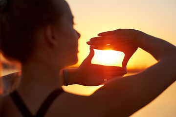 Woman silhouette framing sun with fingers at sunset. Hand shape of a camera across the sky.