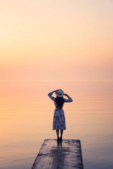 Woman wearing elegant dress with hat standing on the pier over colorful sea water and twilight sky background.