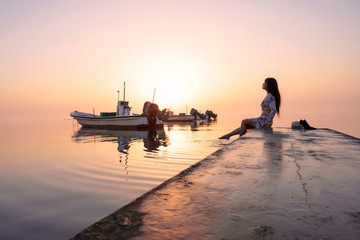 Beautiful Woman sitting on the bridge pier feet in sea water over colorful morning light reflection over sky with boats floatings background