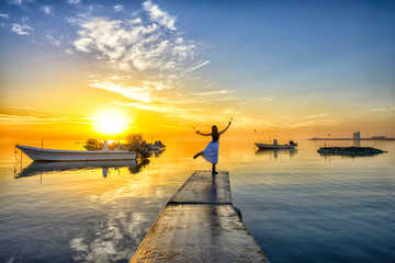 Beautiful sunrise image with boats parking on concrete pier on sea water with movement of woman in white dress dancing with birds flying over colorful sky background, Bahrain.