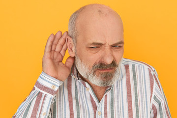 Portrait of frowning frustrated bearded male pensioner in striped shirt keeping hand at his ear,...