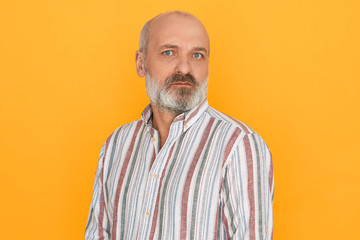 Obraz premium Portrait of handsome European male pensioner with bald head and thick gray beard looking at camera having doubtful suspicious facial expression, not trusting you. Human emotions and reaction