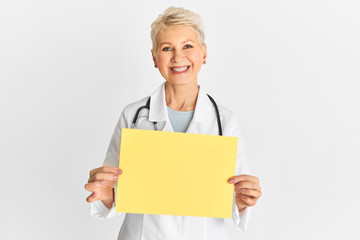 Portrait of cheerful beautiful middle aged female doctor or nurse wearing medical white coat showing blank empty sign board with copy space for text or advertisement, smiling broadly at camera