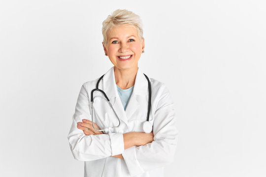 Isolated shotof happy successful mature senior physician wearing medical unifrom and stethoscope having cheerful facial expression, smiling broadly, keeping arms crossed on chest, enjoying her job