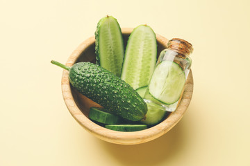 Bottle of essential oil and cucumbers in bowl on color background