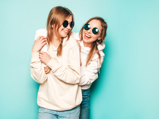 Two young beautiful blond smiling hipster girls in trendy summer hoodie clothes. Sexy carefree women posing near blue wall. Trendy and positive models having fun in sunglasses