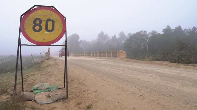 Red Van Drives Past 80km Speed Sign Over A Bridge In The Mist On A Dirt Road In The Countryside