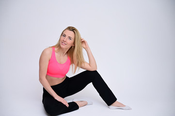 Fototapeta premium Portrait of a pretty fitness blonde girl in a sports uniform on a white background sitting on the floor. A slender Caucasian model poses with a smile.