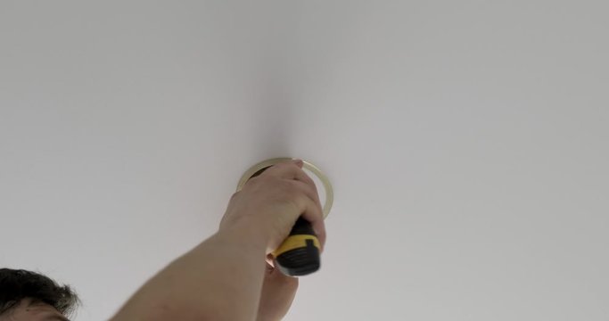 Male electrician carefully cuts circle in new stretch ceiling, cut off round piece with knife, installs lighting in apartment, electrical wiring is located in hole under ceiling, repair in apartment
