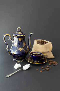 Still life with a beautiful cobalt blue colored vintage porcelain coffee set with golden floral pattern, a silver spoon with sugar cubes and a gunnysack filled with roasted coffee beans.
