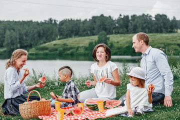 Happy family eating watermelon at a picnic. Picnic in the meadow or park near the lake. Young friends and their children in nature