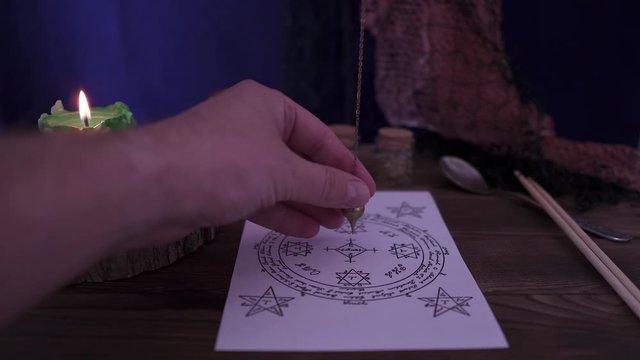 Witchcraft and wizardry. The sorcerer uses the astrological pendulum for Tarot and the occult circle for divination against the background of burning candles and magic potions. Low light, close-up