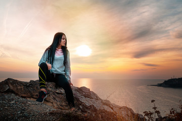 A young woman sitting on the edge of a cliff. In the background, the sea and the sky at sunset.Copy space. Concept of Hiking and outdoor activities