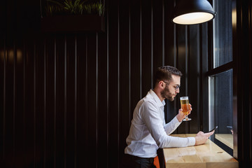 Side view of serious man in shirt sitting in pub next to window, using smart phone for replying...
