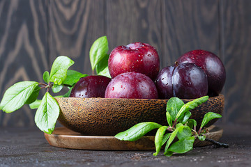 Ripe juicy purple plums with drops in wooden bowl with leaves, dark brown concrete background.