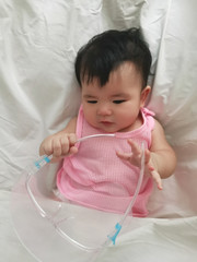 The baby wears a face shield lying on white bedsheet in pink vest.