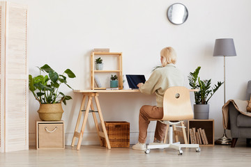 Rear view of woman sitting at the table and working on laptop in the room at home