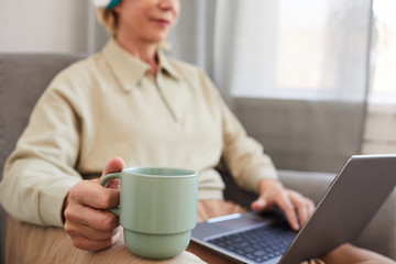 Close-up of woman holding cup of coffee while sitting on sofa with laptop and drinking coffee in the morning