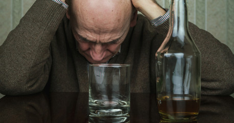 aged alcoholic sits at table with bottle with whiskey and glass holding hands on head