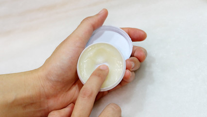 Hand holding a small round container with ointment, and a finger trying to scoop some ointment out.