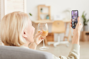 Rear view of mature woman with blonde short hair drinking wine and talking online with her family using her mobile phone