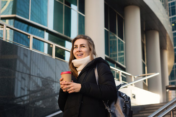 Portrait busy modern pretty woman stands on sunny day. Attractive business lady holding takeaway coffee, using mobile phone. Business woman on background modern business center.