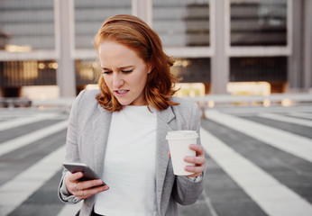 Frustrated young woman in formal clothing reading message on smartphone while holding coffee cup