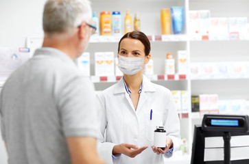 medicine, pharmaceutics and healthcare concept - apothecary wearing face protective medical mask for protection from virus disease showing drug to senior male customer at drugstore
