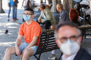 ragazzo con occhiali neri e maglia arancione indossa un mascherina chirurgica seduto  in una panchina nella piazza del centro di una città 