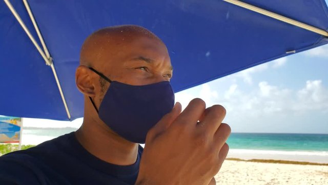 Black African American young man with beard  on beach shows correct way to wear protective face mask in health crisis as beach reopens on travel summer vacation, close up in 4k