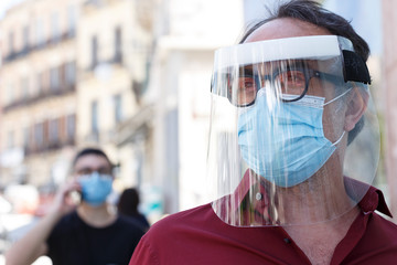 Uomo con mascherina protettiva e maschera in plexiglass professionale  è poggiato sulla vetrina di un negozio in cui si riflette la città