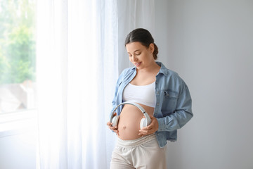 Beautiful pregnant woman putting headphones on her belly at home