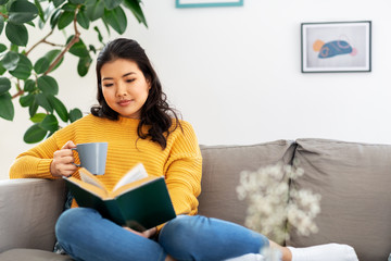 people and leisure concept - happy asian young woman in yellow sweater sitting on sofa, reading...