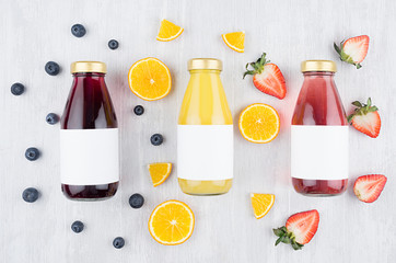 Fresh yellow orange, pink strawberry, violet blueberry juices in glass bottles with blank label on white wood board, mock up for design, advertising.