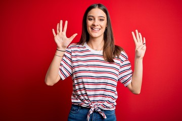 Young beautiful brunette woman wearing casual striped t-shirt standing over red background showing...