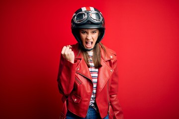 Young beautiful brunette motocyclist woman wearing motorcycle helmet and red jacket angry and mad raising fist frustrated and furious while shouting with anger. Rage and aggressive concept.