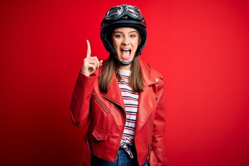 Young beautiful brunette motocyclist woman wearing motorcycle helmet and red jacket pointing finger up with successful idea. Exited and happy. Number one.