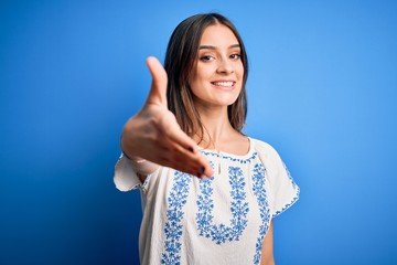 Fototapeta na wymiar Young beautiful brunette woman wearing casual t-shirt standing over blue background smiling friendly offering handshake as greeting and welcoming. Successful business.